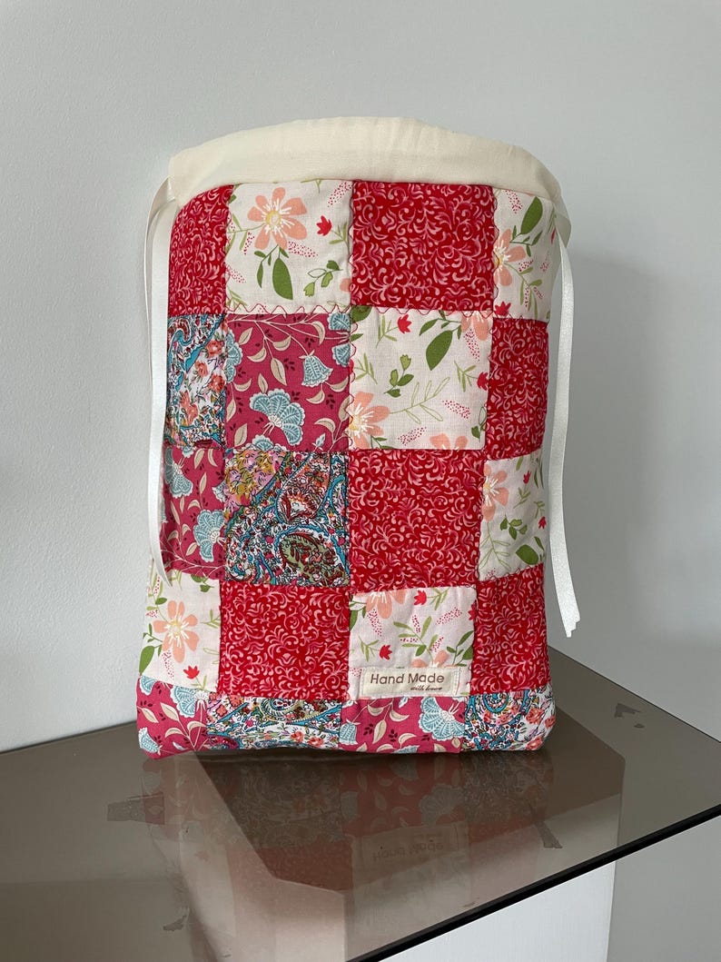 Pu&ograve; includere: Una borsa rettangolare con coulisse caratterizzata da un design patchwork con motivi floreali e paisley rossi, rosa e bianchi. La parte superiore presenta un bordo in tessuto color crema con nastri bianchi. Un'etichetta bianca riporta Hand Made with love.