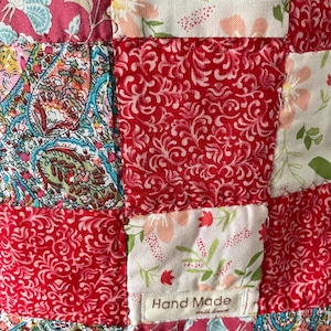 Pu&ograve; includere: Un tessuto patchwork composto da vari quadrati di stoffa nelle tonalit&agrave; del rosso, rosa, bianco e verde acqua. I motivi includono disegni floreali, paisley e fogliame. Una piccola etichetta bianca in tessuto riporta la scritta Hand Made with love.