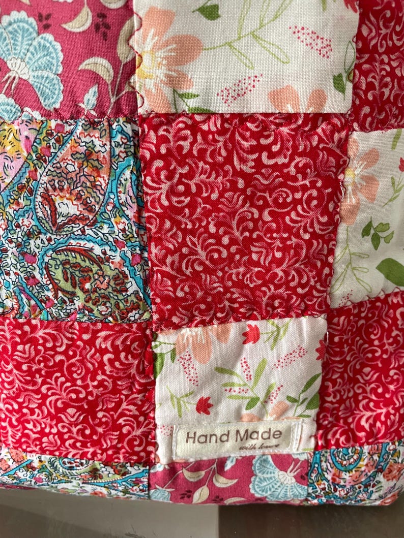 Pu&ograve; includere: Un tessuto patchwork composto da vari quadrati di stoffa nelle tonalit&agrave; del rosso, rosa, bianco e verde acqua. I motivi includono disegni floreali, paisley e fogliame. Una piccola etichetta bianca in tessuto riporta la scritta Hand Made with love.