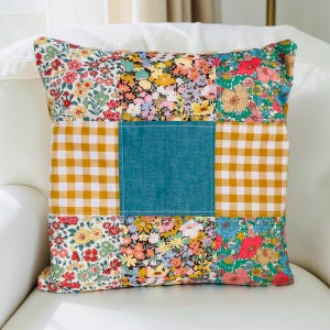 May include: Square patchwork cushion featuring floral and gingham patterns in red, yellow, blue, and green. A teal square is centered on the cushion. The cushion is on a white surface.