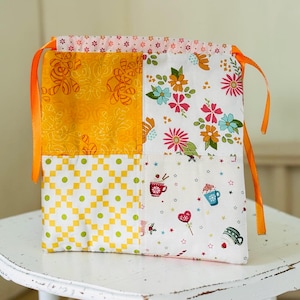May include: A patchwork drawstring bag with orange ribbon ties. The bag features four fabric panels with different patterns: yellow floral, white floral, yellow and green checkered, and a white pattern with coffee cup and heart graphics.