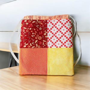 May include: A patchwork drawstring bag with four different fabric patterns. The bag is made of red, orange, yellow, and white fabrics. The bag has a white drawstring and is made of cotton.