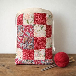 Pu&ograve; includere: Una borsa con coulisse caratterizzata da un design patchwork in tessuti floreali e paisley rossi, rosa e crema. La borsa presenta una parte superiore color crema con lacci in nastro bianco e una piccola etichetta rettangolare con la scritta Hand Made.