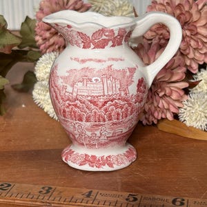 May include: A white ceramic creamer pitcher featuring a red transferware pattern of a castle and figures, with a scalloped rim and handle. A wooden ruler placed in front shows the pitcher stands approximately 4.5 inches tall at the spout.