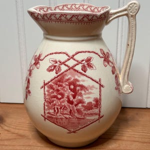 May include: A cream-colored ceramic pitcher featuring a red transferware pattern. The design includes a central hexagonal landscape scene labeled Magna Charta Island, surrounded by oak leaves and acorns, with a woven border at the rim.