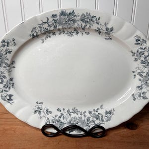 May include: An oval ceramic serving platter featuring a white base with a decorative border of dark gray floral patterns. The platter has a scalloped edge and a smooth, glossy finish, showcasing intricate botanical illustrations of flowers and leaves.