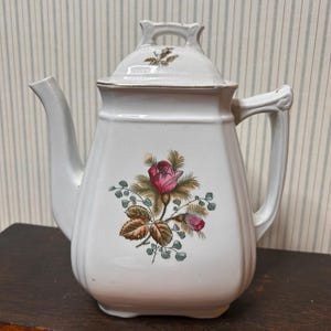May include: A white ceramic teapot with a square-shaped body and a matching lid. The front features a floral decal of a pink rose with green leaves and small foliage. The teapot has a curved spout, a sturdy handle, and a gold-toned rim detail.