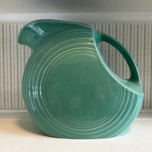 May include: A round, teal-colored ceramic pitcher featuring a series of concentric circular embossed rings on its side, a curved handle, and a wide pouring spout. The surface has a smooth, glossy finish with a consistent solid color throughout.