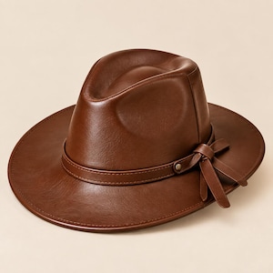 Può includere: Un cappello fedora marrone con una finitura liscia simile alla pelle. Il cappello presenta una corona strutturata con una piega centrale e una tesa larga. Una fascia marrone abbinata circonda la base della corona, rifinita con un fiocco decorativo sul lato.