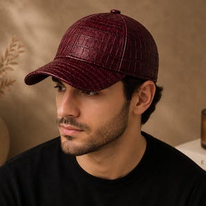 Puede incluir: Una gorra de béisbol de color burdeos intenso con un patrón texturizado de piel de cocodrilo. La gorra tiene una corona estructurada, una visera curva y un pequeño botón en la parte superior central. El material tiene un acabado brillante que capta la luz, resaltando las escamas en relieve en toda la superficie.