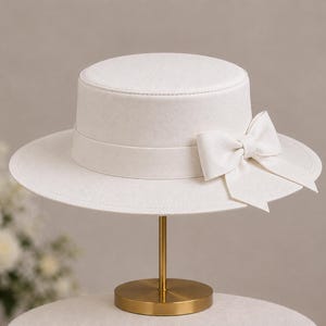 May include: A white flat-top hat with a wide brim and a matching white ribbon band featuring a decorative bow on the side. The hat is displayed on a gold-coloured metal stand with a circular base, showcasing a clean and structured design.