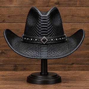 May include: A black cowboy hat with a textured snakeskin-like pattern. The hat features a black band adorned with silver-toned studs and a central diamond-shaped metal emblem. The brim is curved upwards on both sides, and the crown has a deep centre crease.