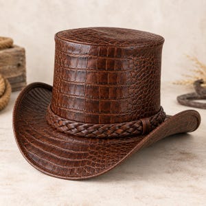 Puede incluir: Un sombrero de copa marrón con un patrón de textura de piel de cocodrilo en toda su superficie. El sombrero incluye un ala ancha y curva y una banda trenzada decorativa que rodea la base de la copa, proporcionando un aspecto estructurado y uniforme.