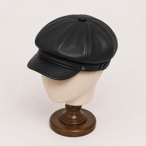 Puede incluir: Una gorra de estilo boina en cuero negro con una corona estructurada de ocho paneles, un botón central y una visera corta y curva. Una tira fina de cuero con una pequeña hebilla metálica y un remache lateral se sitúa sobre la base de la corona.