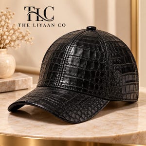 Pode incluir: Um boné de beisebol preto com um padrão de textura de pele de crocodilo brilhante. O boné apresenta uma coroa estruturada e uma aba curva. O texto TLC THE LIYAAN CO está impresso em preto acima do boné no fundo.