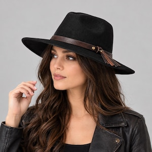 Könnte beinhalten: Ein schwarzer Fedora-Hut mit breiter Krempe und einem dunkelbraunen Lederband um die Krone. Das Band ist mit einem kleinen goldfarbenen Metallniet und einer dekorativen braunen Lederquaste verziert, die an der Seite des Hutes herabhängt.