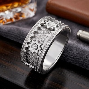 May include: A silver-toned ring featuring a wide band with a mechanical gear design. The surface is encrusted with numerous small, clear, sparkling stones, and each gear element is accented with a single, larger, centered clear stone.
