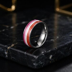 May include: A polished silver-toned metal ring featuring a central inlay with horizontal stripes in shades of dark orange, light orange, white, light pink, and dark pink. The band has a smooth, reflective finish and a rounded profile.
