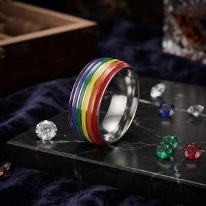 May include: A silver-toned metal ring featuring a central band with horizontal stripes in red, orange, yellow, green, blue, and purple enamel. The ring has a polished finish and is displayed alongside several loose, faceted, multi-coloured gemstones.