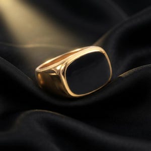 May include: A gold-toned signet ring featuring a smooth, polished surface with a large, rectangular black inlay centered on the face. The band is thick and solid, showcasing a reflective metallic finish that contrasts with the deep black center.