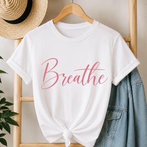 Peut inclure: Un t-shirt blanc à manches courtes avec le mot Breathe imprimé en écriture cursive rose au centre. Le vêtement présente une encolure ronde et est suspendu à un cintre en bois avec un nœud noué à l'ourlet inférieur du t-shirt.