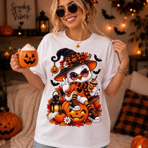 Peut inclure: Un t-shirt blanc avec un motif représentant un hibou de dessin animé portant un chapeau de sorcière, une écharpe et des baskets, assis sur une citrouille d'Halloween. Le design inclut des feuilles d'automne, des chauves-souris et une lanterne.