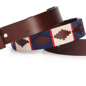 Pode incluir: Um cinto de couro castanho com uma secção de tecido entrançado com um padrão de diamante repetitivo em azul-marinho, creme e vermelho. O cinto inclui uma tira de couro castanho lisa, uma fivela de metal e uma presilha de couro.