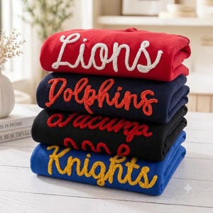 May include: A stack of four folded sweatshirts in red, navy blue, black, and royal blue. Each sweatshirt features textured, raised chenille-style lettering in a cursive font. The text reads Lions in white, Dolphins in orange, Jaguars in red, and Knights in yellow.