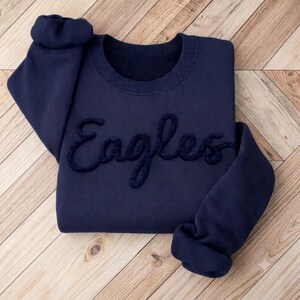 May include: A dark navy blue long-sleeve sweatshirt featuring the word Eagles written in a raised, textured, cursive script across the chest. The garment has a crew neckline and ribbed cuffs, showcasing a solid colour design with matching embroidery.