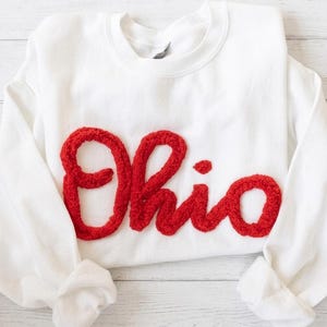 May include: A white crewneck sweatshirt featuring the word Ohio written in large, textured, red chenille-style script lettering across the chest. The garment has long sleeves with ribbed cuffs and a classic round neckline, showcasing a clean design.