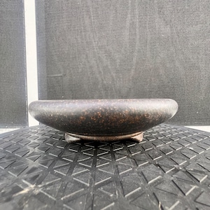 May include: A low-profile, circular ceramic bowl with a dark brown, speckled, matte finish. The bowl features a thick, rounded rim and is supported by three small, integrated feet that elevate the base slightly above the surface.