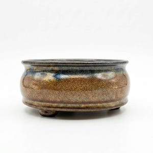 May include: A round ceramic bonsai pot with a low profile and three small feet. The exterior features a textured, mottled brown glaze on the lower body, transitioning to a smooth, dark charcoal and deep blue glaze around the upper rim.
