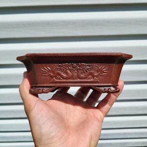 May include: A rectangular terracotta-colored bonsai pot featuring an embossed relief of two dragons facing each other on the front side. The vessel has a wide, flared rim, a flat base, and four small, decorative feet at the corners.