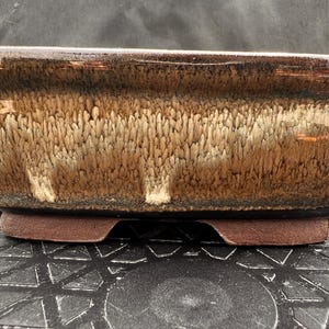 May include: A rectangular ceramic bonsai pot featuring a glossy, multi-toned glaze that transitions from a dark brown rim to a lighter, textured cream and tan drip pattern. The base is made of unglazed, matte brown clay with small feet.