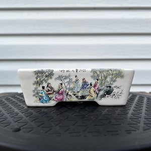 May include: A rectangular white ceramic planter featuring a detailed hand-painted scene of seven figures in traditional robes gathered among trees and rocks. The artwork includes black Chinese calligraphy characters along the top edge of the vessel.