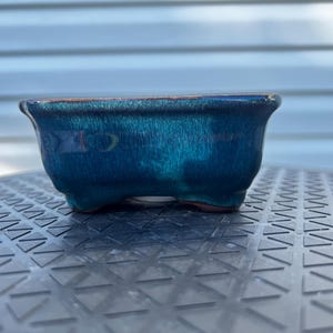 May include: A rectangular ceramic bonsai pot with a glossy, deep teal and blue glaze. The vessel features a flared rim, slightly curved sides, and four small, integrated feet at the base, showcasing a smooth, reflective finish throughout.