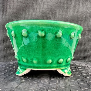 May include: A vibrant emerald green ceramic planter featuring a glossy crackle glaze finish. The bowl-shaped vessel is adorned with two horizontal rows of raised, rounded studs and is supported by three small, curved feet with white undersides.