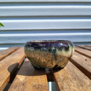 May include: A small, round ceramic planter with three short feet. The glaze features a deep, dark purple rim that transitions into a mottled, earthy tan and light green body with a glossy, smooth finish and a wide, open circular top.
