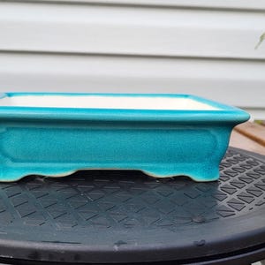 May include: A rectangular ceramic bonsai pot with a vibrant turquoise glazed exterior and a smooth white interior. The pot features a wide, flat rim, slightly tapered sides with a subtle recessed panel design, and four small, sturdy feet at the base.