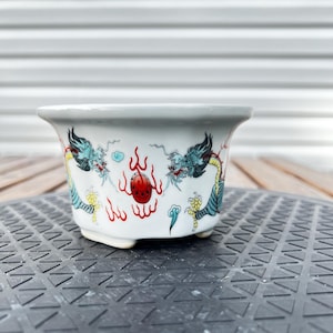 May include: A white ceramic hexagonal planter featuring a hand-painted design of two blue dragons facing a central red flaming pearl. The vessel has a flared rim, a smooth glossy finish, and small feet at the base, showcasing intricate details.