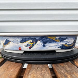 May include: A shallow, oval ceramic bonsai pot featuring a hand-painted design of a long, winding blue dragon with yellow accents. The dragon is depicted amidst white clouds against a light blue background, with a small red orb near its head.