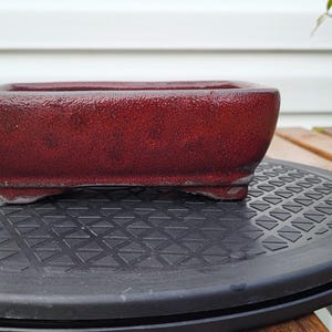 May include: A rectangular bonsai pot with a deep red, textured, and mottled finish. The container features slightly rounded corners, a thick rim, and small, integrated feet at the base, providing a stable foundation for plant cultivation.