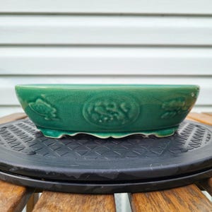 May include: A shallow, round, emerald green ceramic bonsai pot with a glossy, crackled glaze finish. The exterior features embossed, circular decorative patterns along the sides and is supported by small, integrated feet at the base.