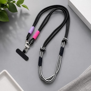 May include: A black cord lanyard featuring a silver metal swivel clasp and a black rectangular attachment tab. The cord includes decorative accents in lavender and hot pink, along with sections of black and white patterned rope with dark knots.