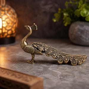 May include: A metallic gold-toned peacock figurine featuring intricate embossed feather detailing on its body and a long, fanned-out tail with circular eye patterns. The bird stands on two small feet with its head turned slightly to the left.