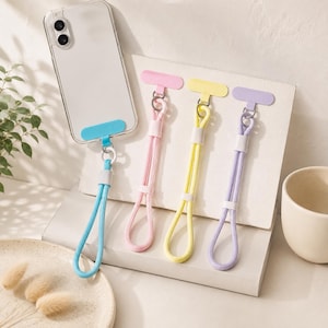 May include: A clear smartphone case with a blue adhesive tether tab and a matching blue wrist strap, alongside three additional wrist straps in pastel pink, yellow, and purple, each featuring a flat adhesive tab and a metal ring connector.