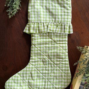 May include: A quilted Christmas stocking featuring a light green and white gingham check pattern. The stocking includes a ruffled trim detail at the top cuff and a small fabric loop for hanging. The main body of the stocking has a diamond-stitched pattern.