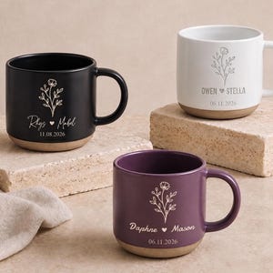 Puede incluir: Tres tazas de cerámica en negro, blanco y morado, cada una con un dibujo floral, dos nombres separados por un corazón y una fecha. Las tazas tienen un acabado liso en la parte superior y una base sin esmaltar de color beige en la parte inferior.