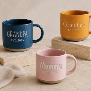 May include: Three ceramic mugs in blue, yellow, and pink. The blue mug features the text GRANDPA EST. 2025, the yellow mug features Grandma EST. 2025, and the pink mug features Mommy EST. 2026. Each mug has a handle and a light-colored base.