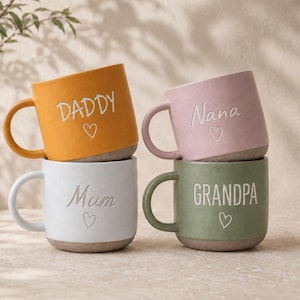 Puede incluir: Cuatro tazas de cerámica en amarillo mostaza, rosa suave, blanco y verde salvia. Cada taza tiene un asa, una base texturizada y una palabra grabada: DADDY, Nana, Mum o GRANDPA, acompañada de un pequeño símbolo de corazón bajo el texto.